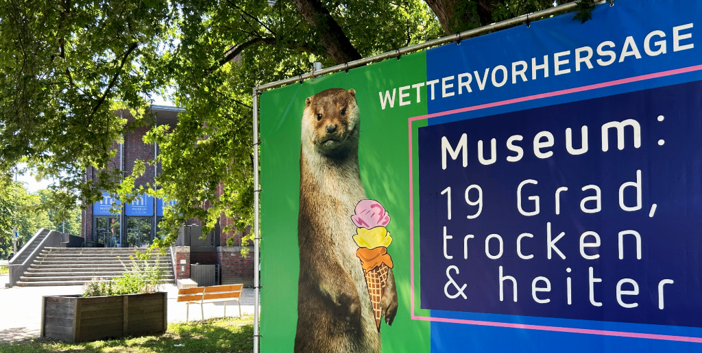 Banner vor dem Museum Lüneburg mit der Aufschrift "Wettervorhersage: Museum: 19 Grad, trocken & heiter.", daneben ist ein Fischotter abgebildet, der eine Eistüte mit drei Kugeln hält.