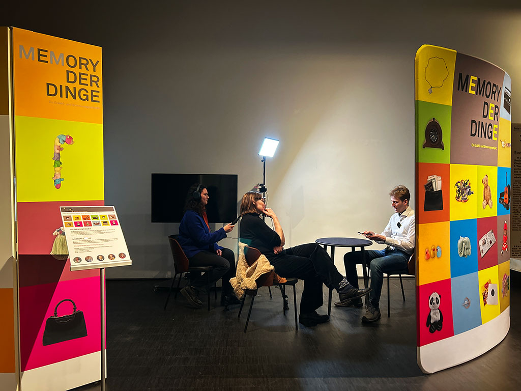 Interviewszene im Memory-Studio im Museum Lüneburg. Eine Person sitzt an einem Tisch, hält ein Objekt in der Hand und erzählt dessen Geschichte. Auf der anderen Seite hört die Interviewerin interessiert zu und eine zweite Frau filmt das Interview.