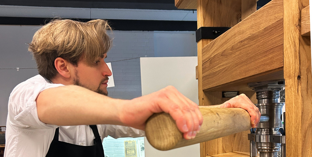 Startseite_Schaudrucken_Lueneburg Ein Mann mit Schürze drückt einen Hebel an einer Druckerpresse aus Holz